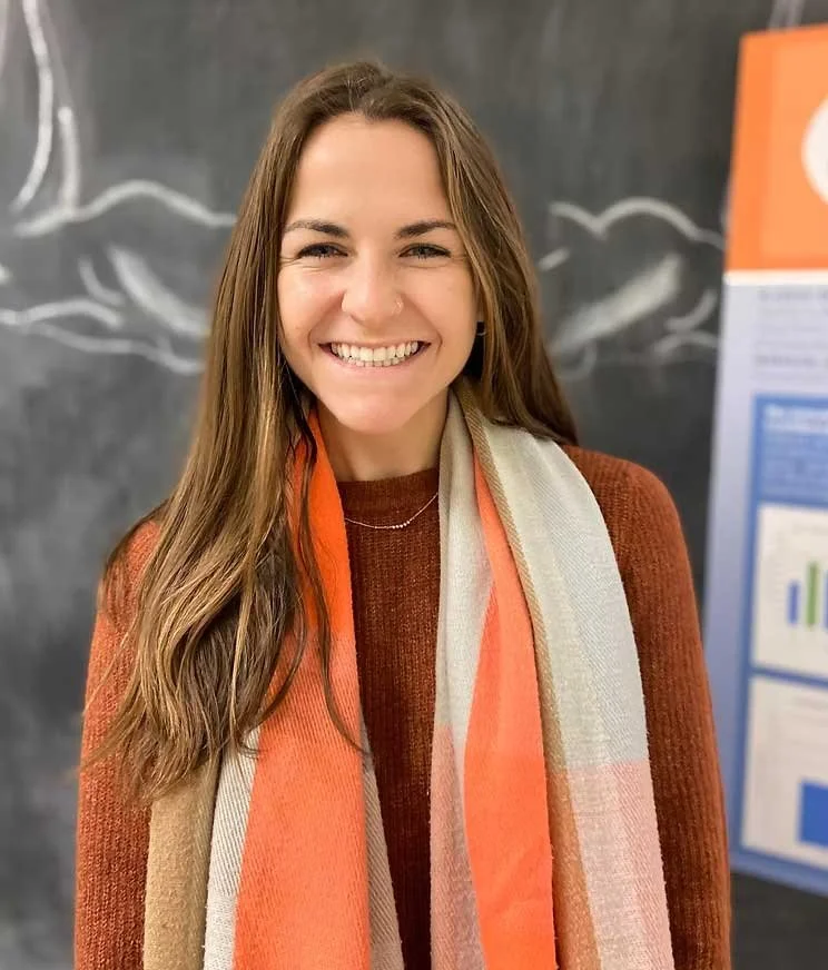 A young woman with long brown hair smiling, standing in front of a blackboard with drawings, wearing an orange-brown sweater and a multicolored scarf.