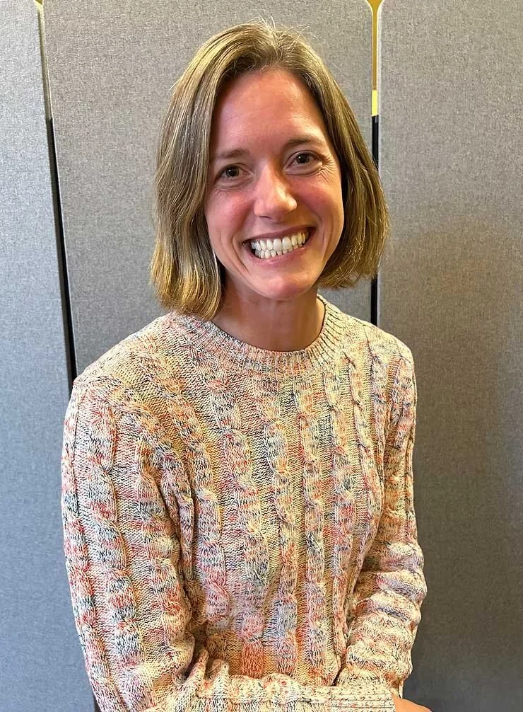 A woman with shoulder-length light brown hair smiling at the camera, wearing a multicolored knitted sweater, standing indoors with a gray partition in the background.