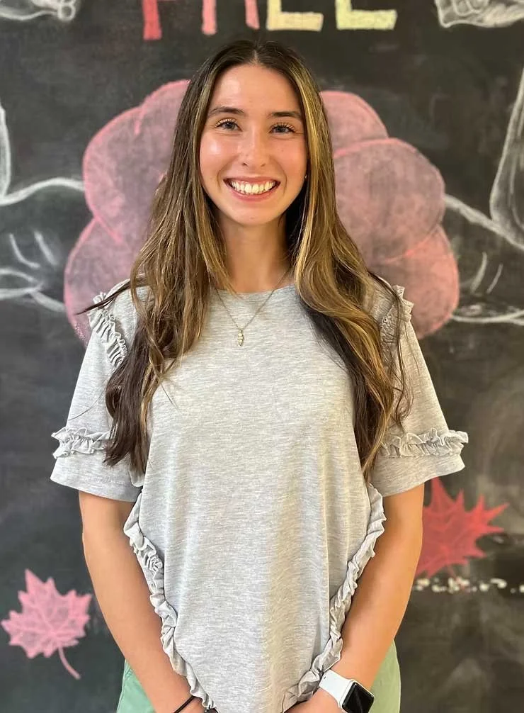 A young woman with long brown hair smiling, wearing a light gray ruffled T-shirt, a necklace, and a white smartwatch, standing in front of a chalkboard decorated with pink chalk flowers and the word 'Spring' written at the top.
