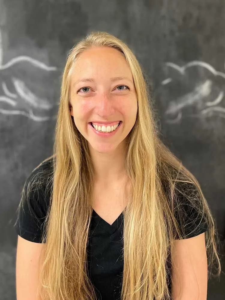 A young woman with long blonde hair smiling in front of a chalkboard background with white chalk drawings of clouds.
