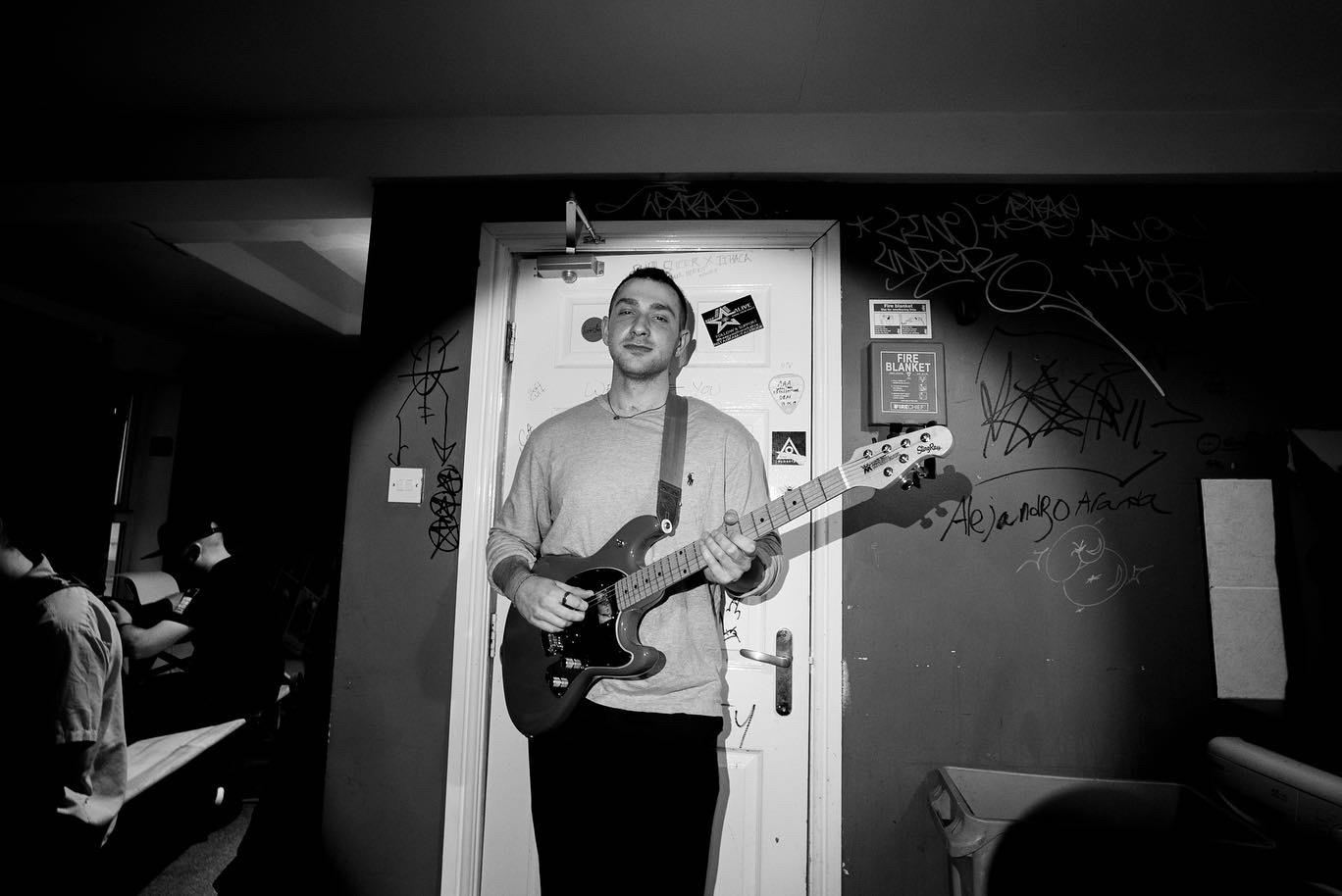 A young man with short hair holding an electric guitar, standing in front of a graffiti-covered wall and a door.