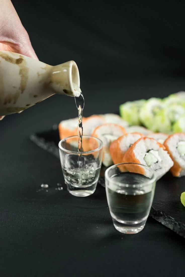 A hand pouring sake into a clear glass shot glass with sushi rolls in the background on a black surface.