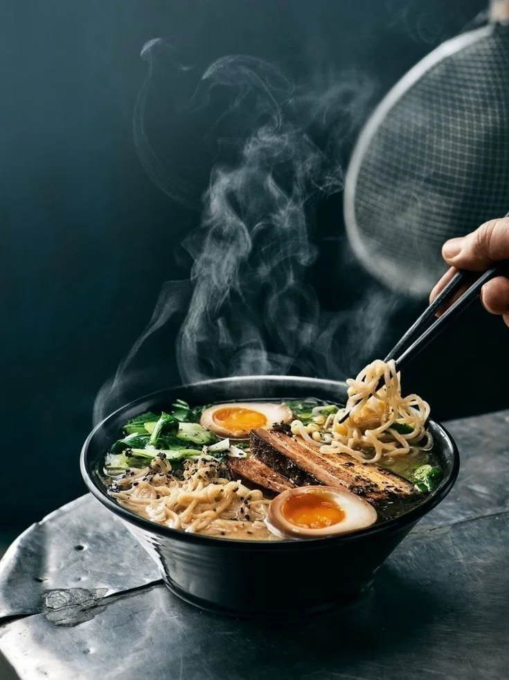 A steaming bowl of ramen with soft-boiled eggs, sliced pork, green vegetables, and noodles, with chopsticks lifting some noodles.