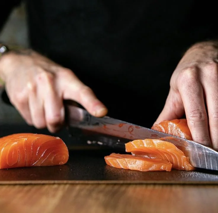 Someone slicing fresh salmon fillet on a black cutting board with a knife.