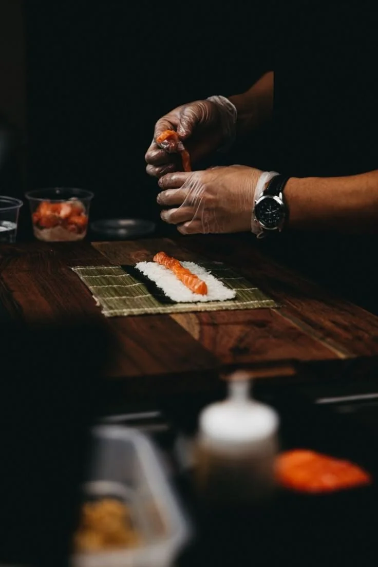 A person wearing gloves and a wristwatch is preparing sushi, placing raw salmon on a bed of rice on a bamboo mat.