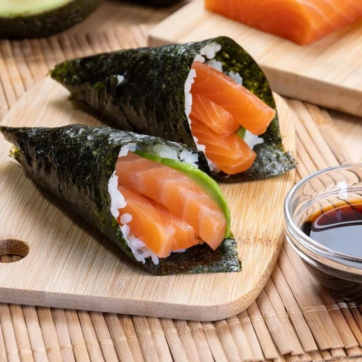 Two salmon sushi hand rolls with nori wrapping, served on a wooden cutting board with soy sauce in a small bowl.