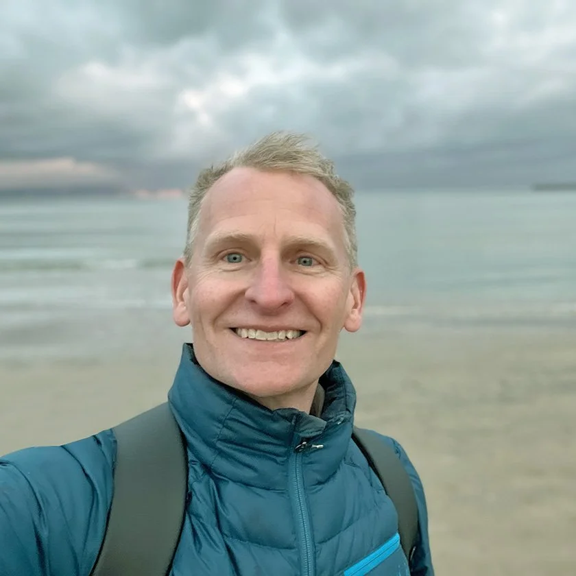A smiling man with short blonde hair and blue eyes taking a selfie on a beach with cloudy skies in the background. He is wearing a blue jacket and has a backpack.