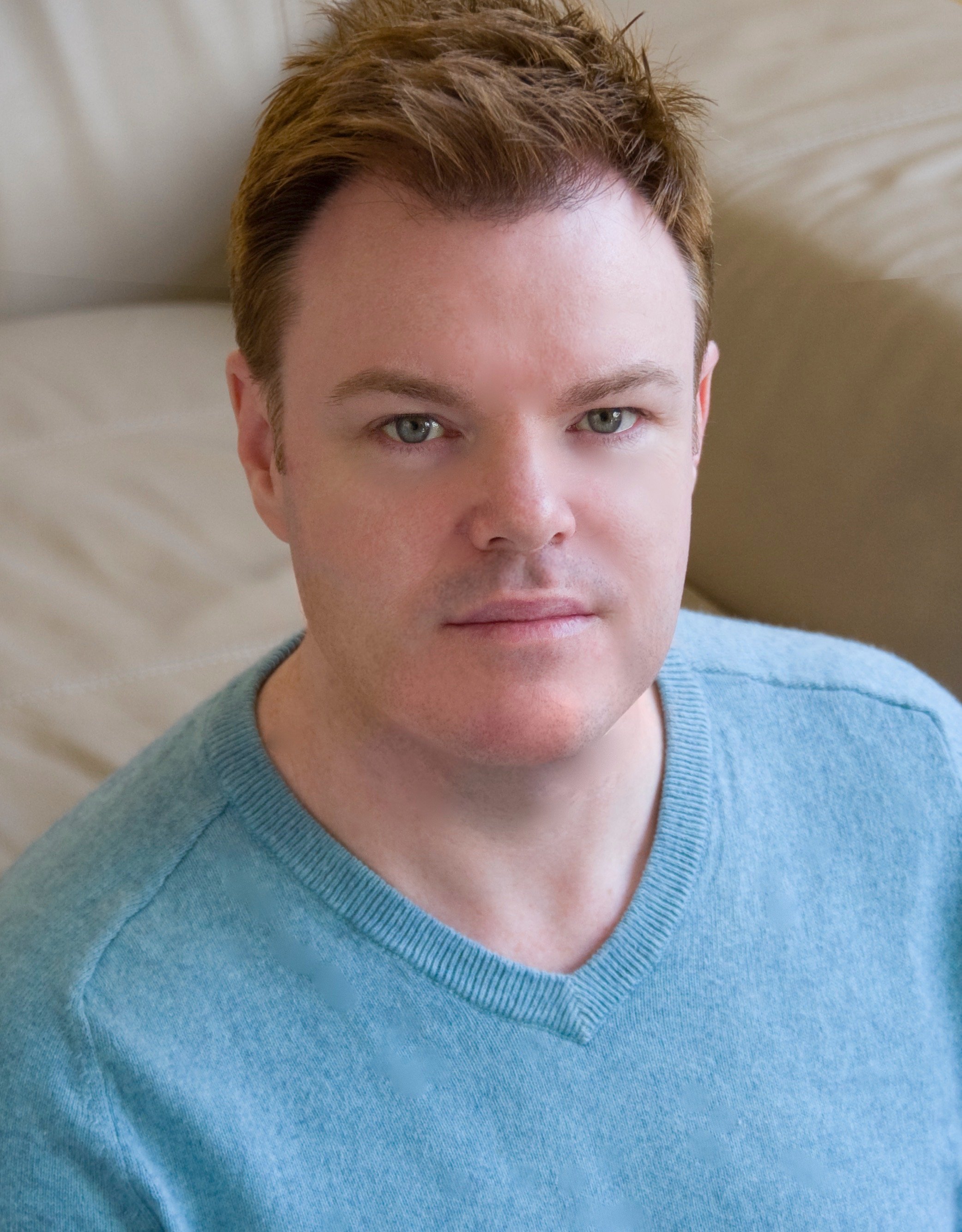 A young man with light skin and reddish-brown hair, wearing a blue V-neck shirt, is looking directly into the camera with a neutral expression. He is sitting against a beige headboard or cushion.