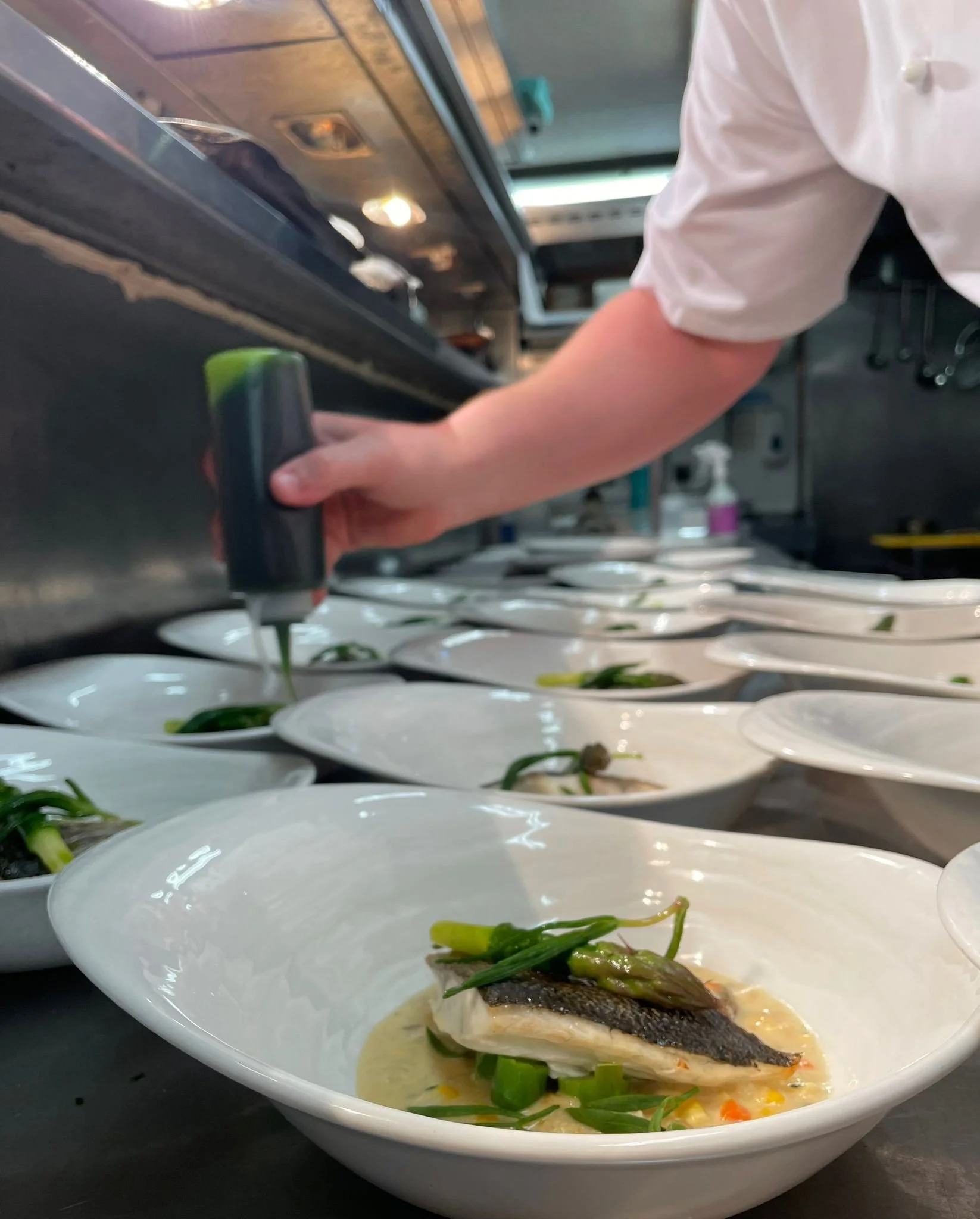 Chef garnishing a plated dish with sauce in a professional kitchen, with multiple plates of the same dish lined up on a counter.