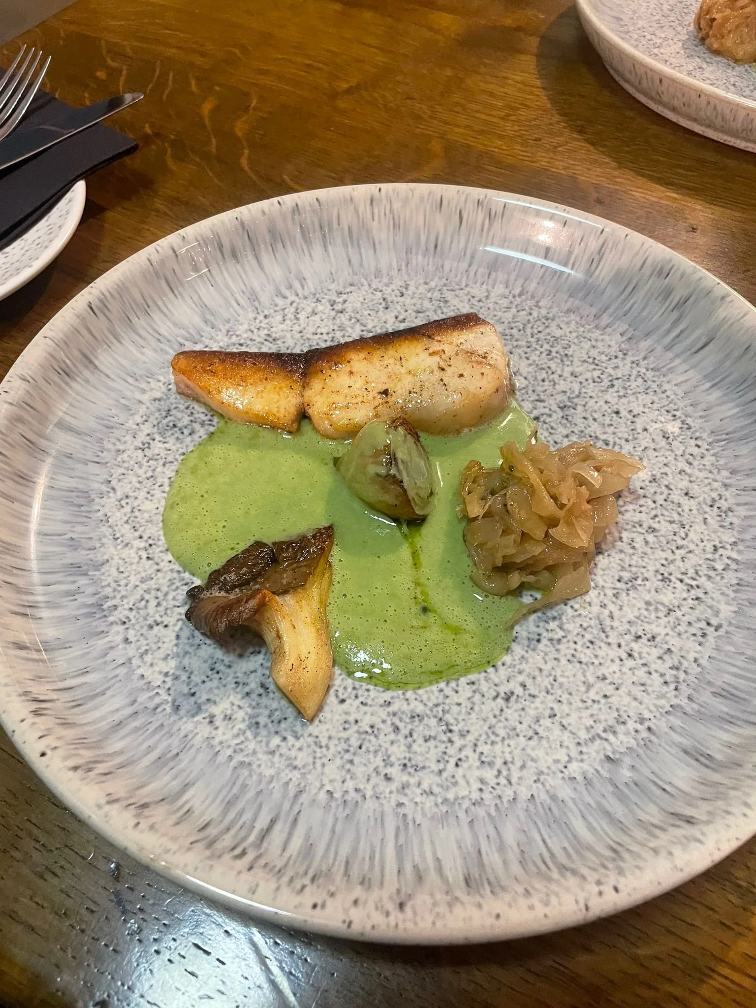 A plated dish with seared fish, mushrooms, cabbage, and green sauce on a patterned ceramic plate.