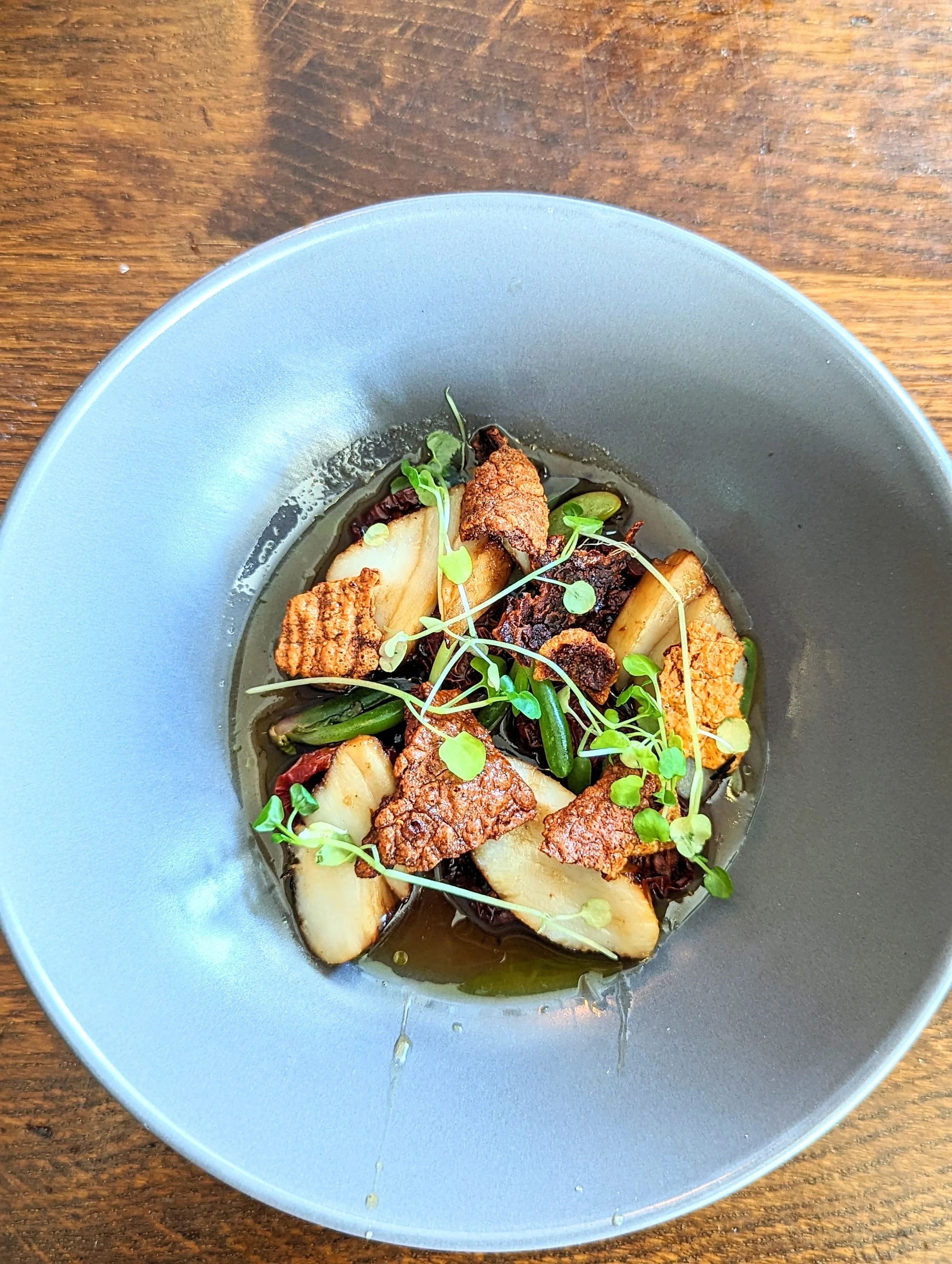 A gray bowl containing a gourmet dish with fingerling potatoes, microgreens, crispy pork belly, and a light broth on a wooden table.