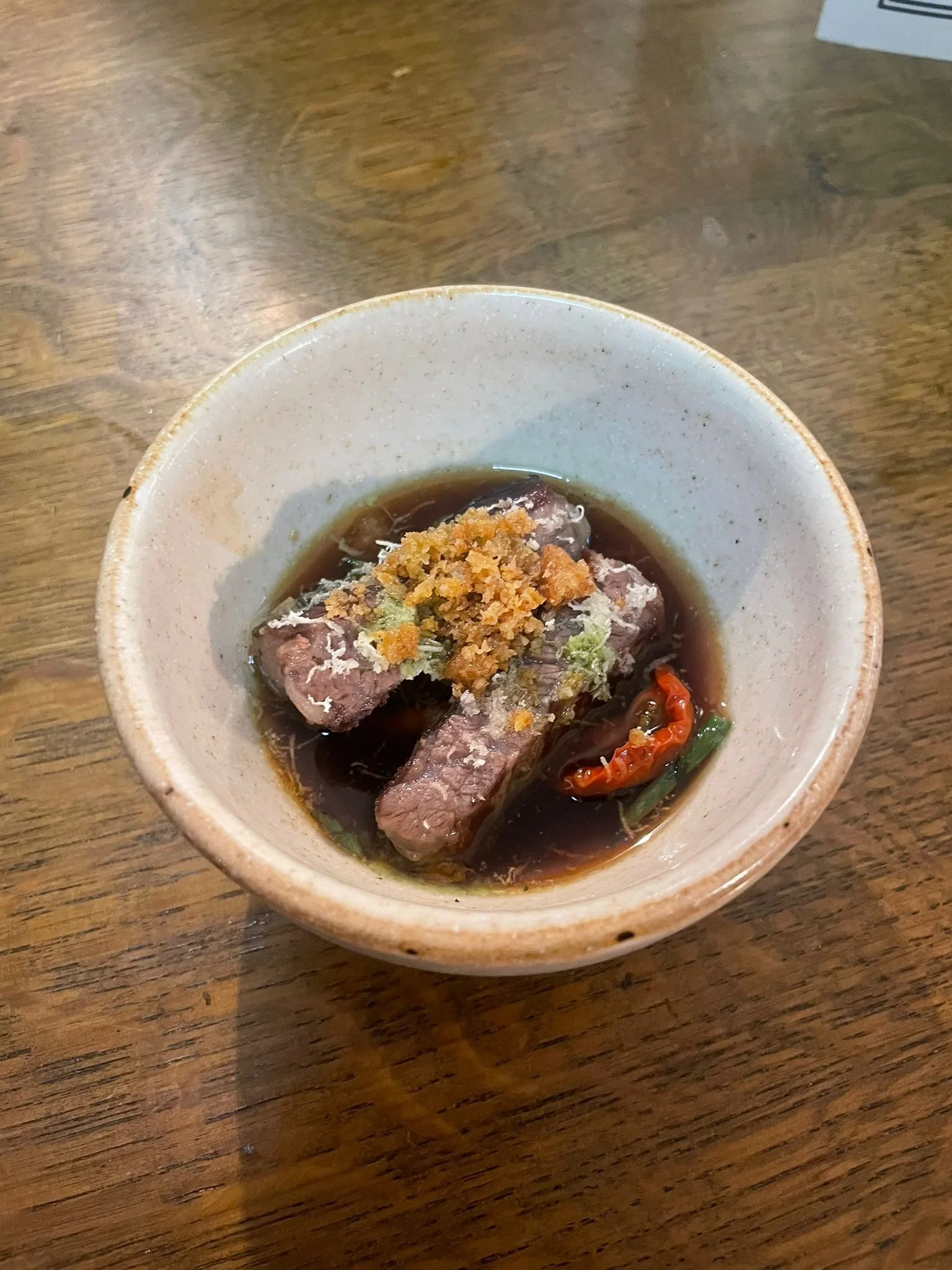 A ceramic bowl containing slices of cooked beef, topped with grated cheese, in a soy-based sauce with a red chili and green vegetables, placed on a wooden table.