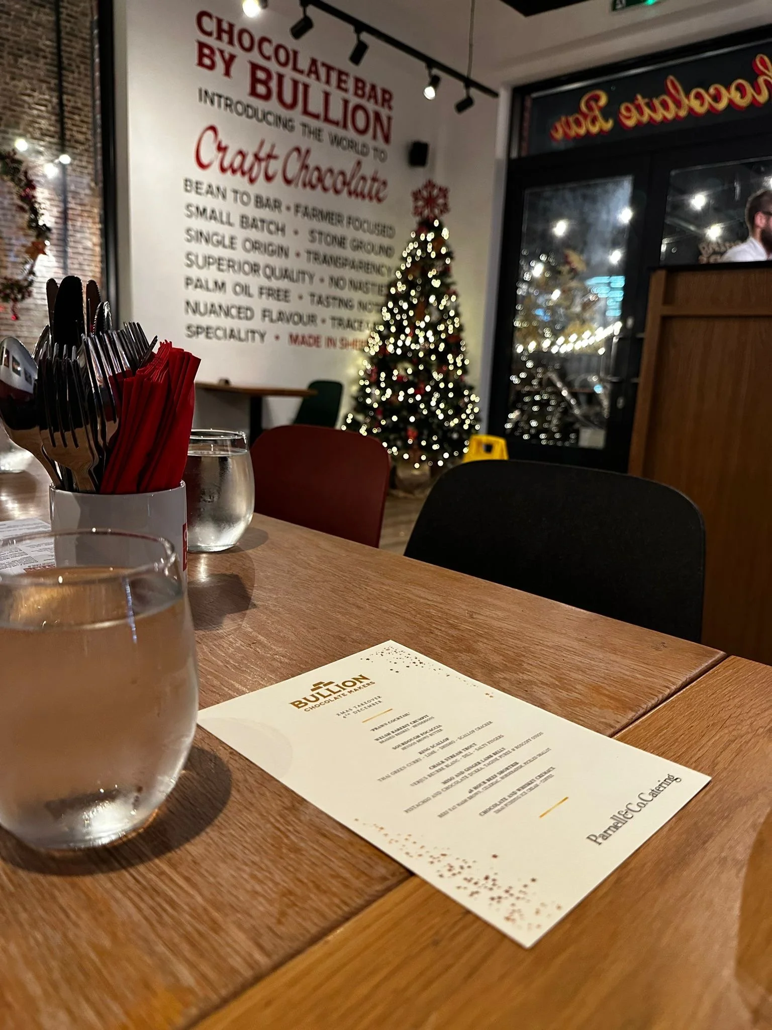 A wooden table with a glass of water, a menu, and utensils wrapped in red napkins inside a restaurant decorated for Christmas, with a Christmas tree and a large white wall with red text in the background.