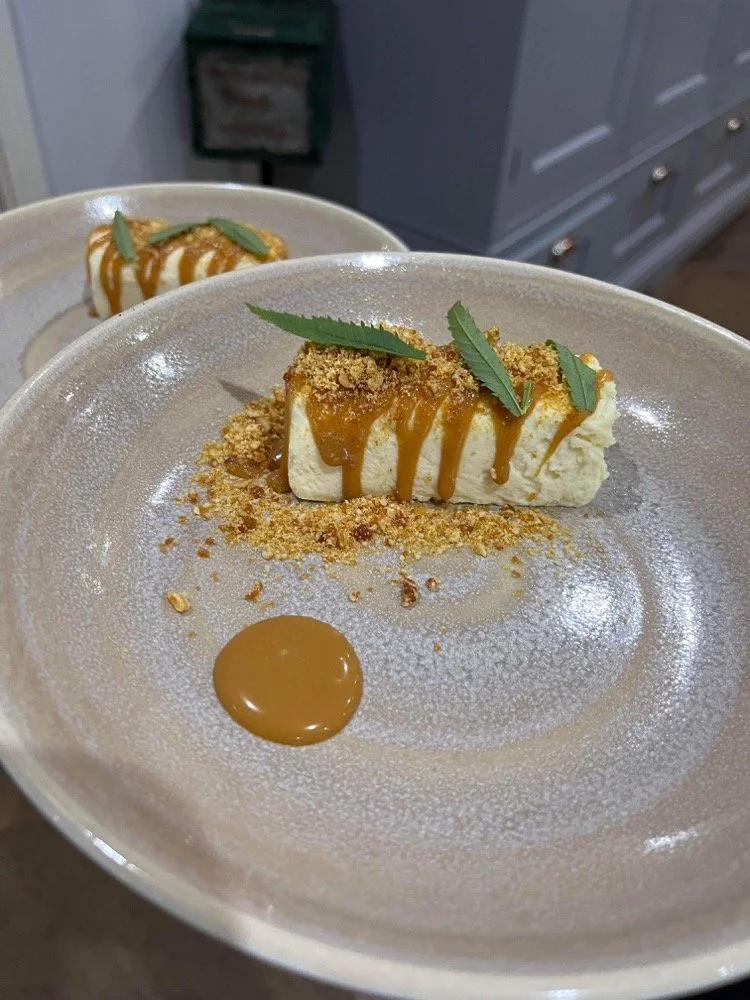 A slice of cheesecake topped with caramel sauce, crushed cookies, and green leaves, served on a plate with a dollop of caramel sauce.
