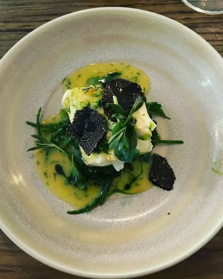 A plated gourmet dish with a piece of white fish, green vegetables, black truffle slices, and a yellow sauce with herbs.