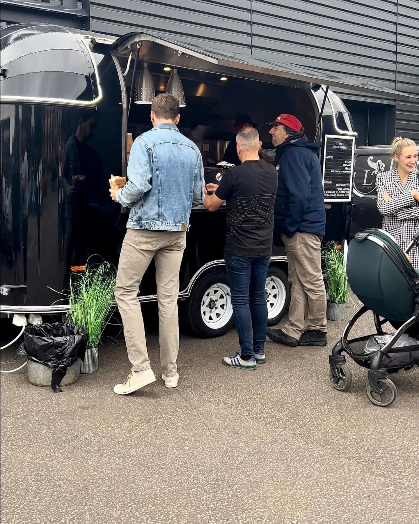 People ordering food at a black food truck with a menu sign, a woman smiling on the right, and a stroller nearby.