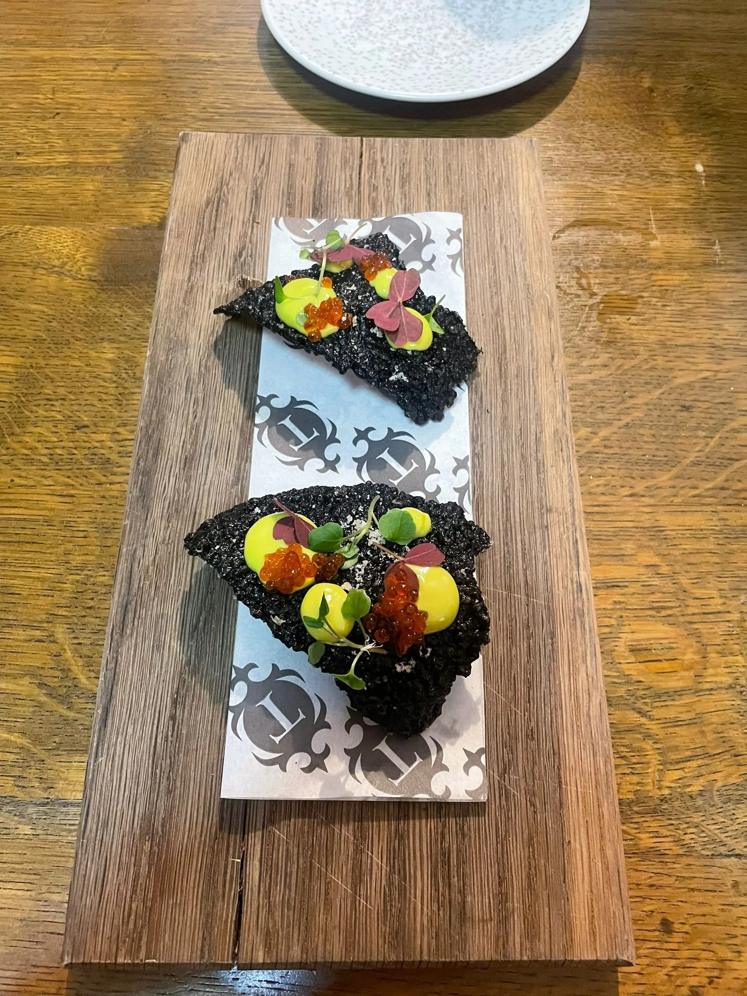 Two pieces of black crispy-textured appetizer topped with yellow sauce, red fish roe, microgreens, and edible purple and green leaves, served on a decorative paper on a wooden platter.