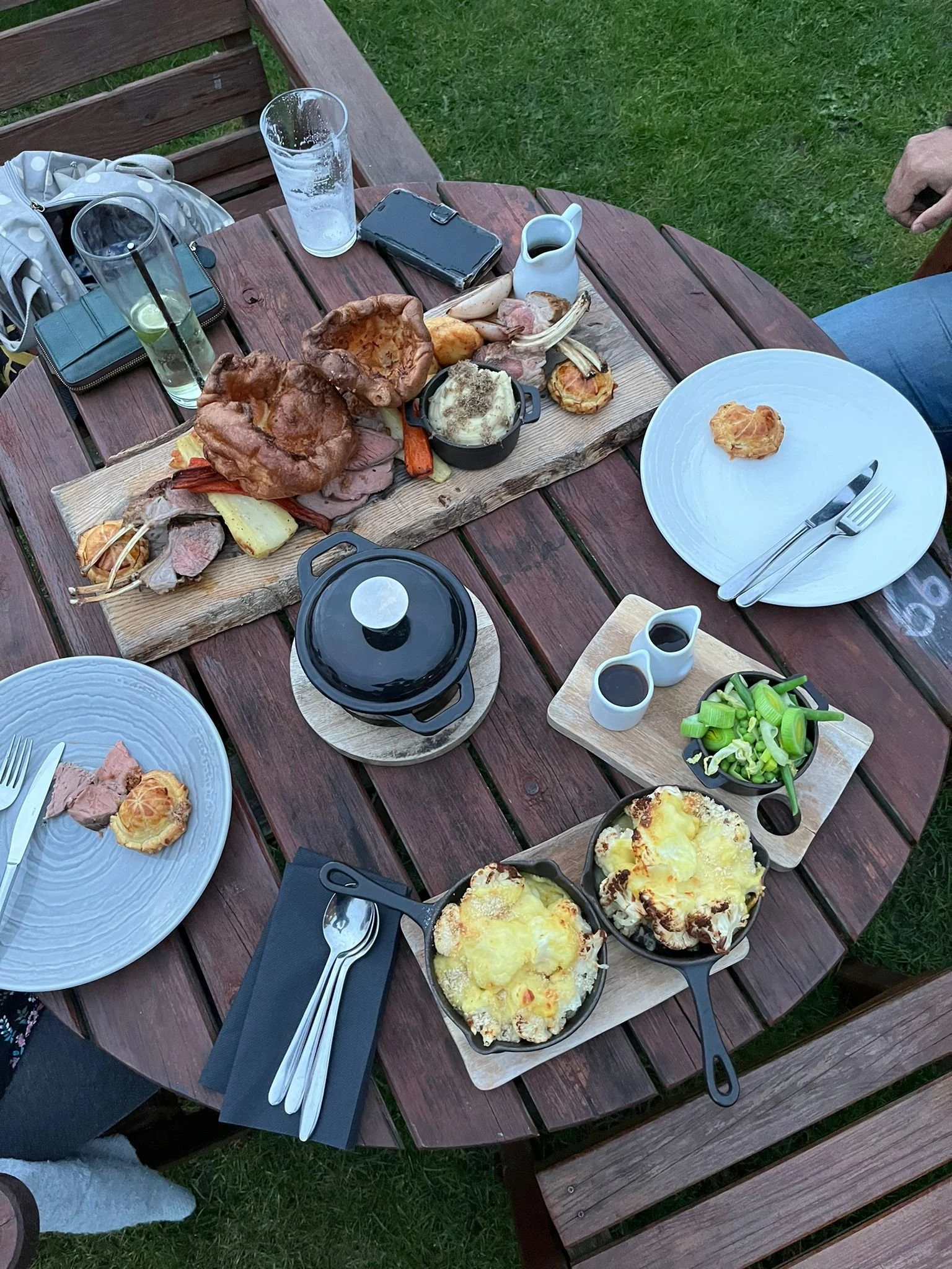 A wooden outdoor table set with food including a platter of roast meats and vegetables, small dishes of sauces, a bowl of chopped green onions, two small cast iron skillets of cheesy mashed potatoes, and empty plates with cutlery, with glasses and pe