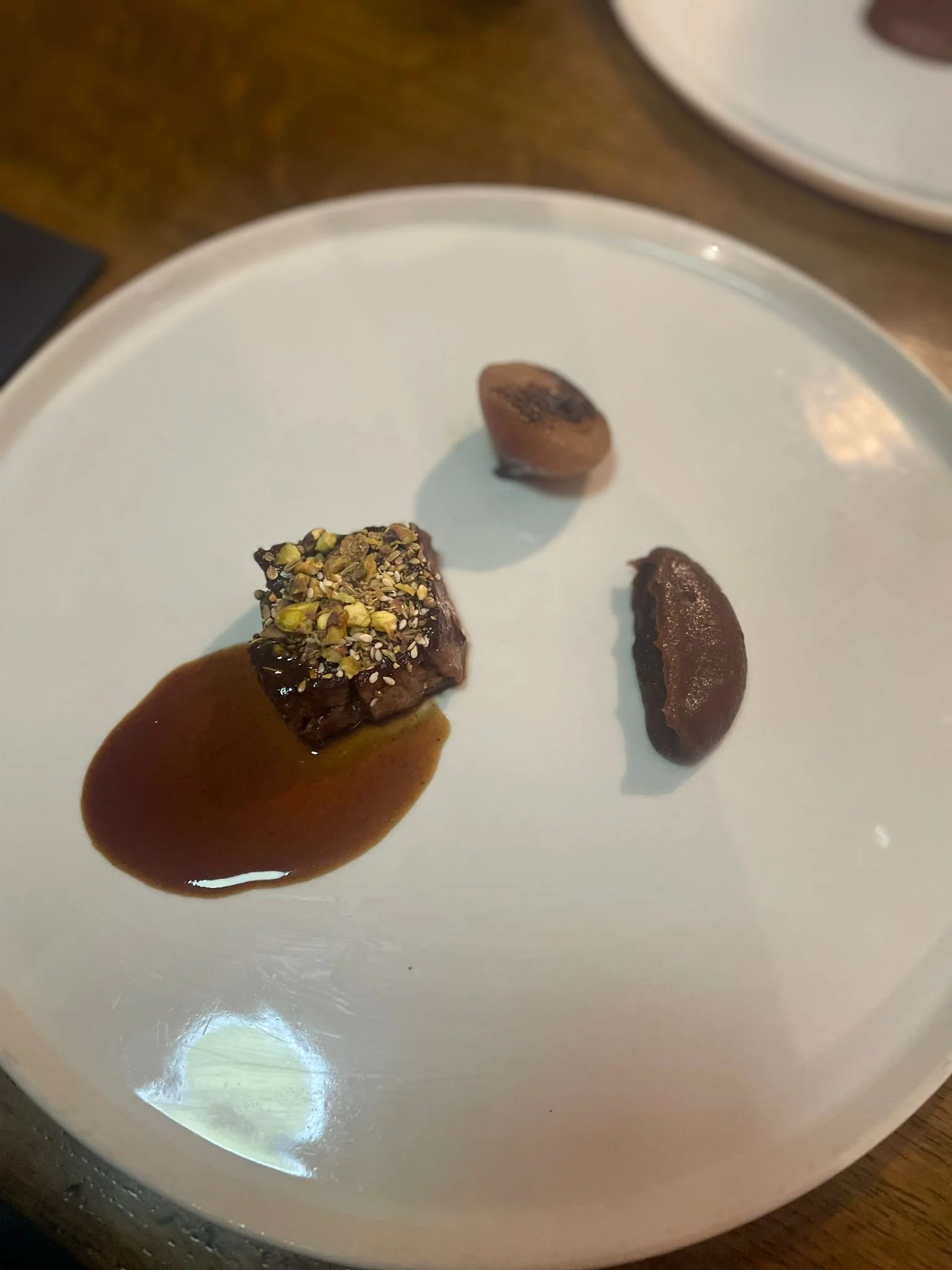 A white plate holding three small pieces of food: a piece of beef topped with chopped pistachios and sauce, and two smaller pieces of chocolate mousse, one with a slightly burnt top.