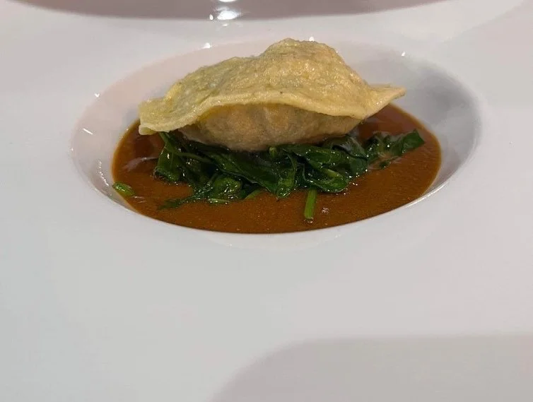 A plated dish featuring a dumpling on a bed of greens, with sauce underneath, served on a white plate.