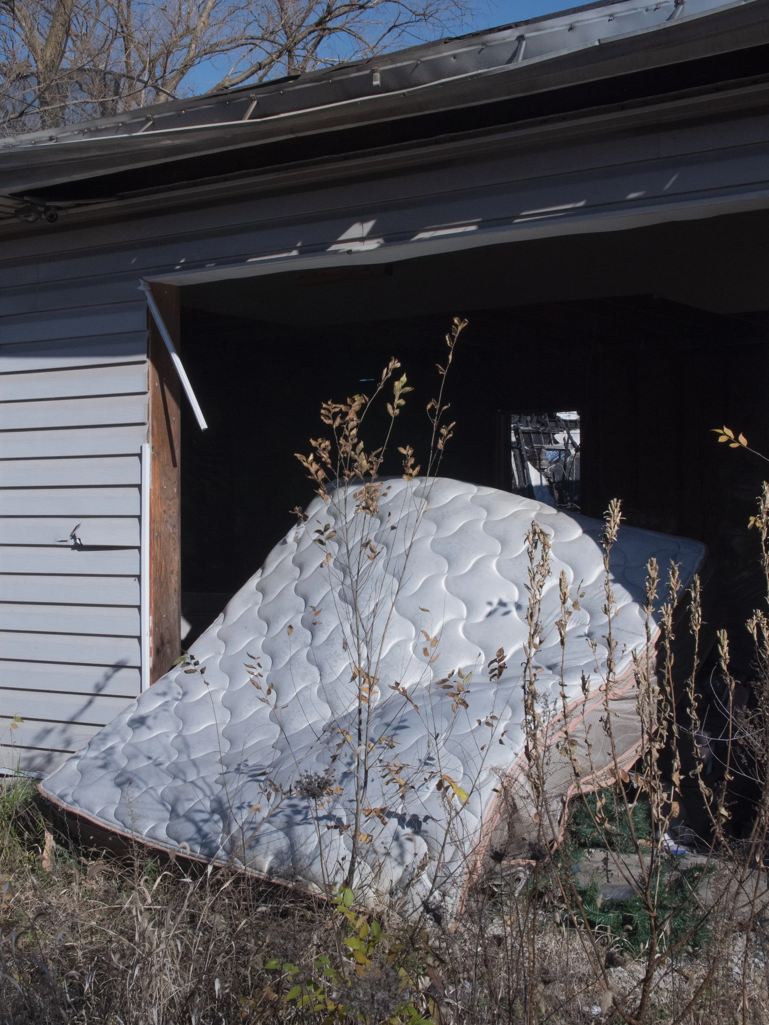 Overgrown yard with an abandoned mattress and debris inside a partially open garage.
