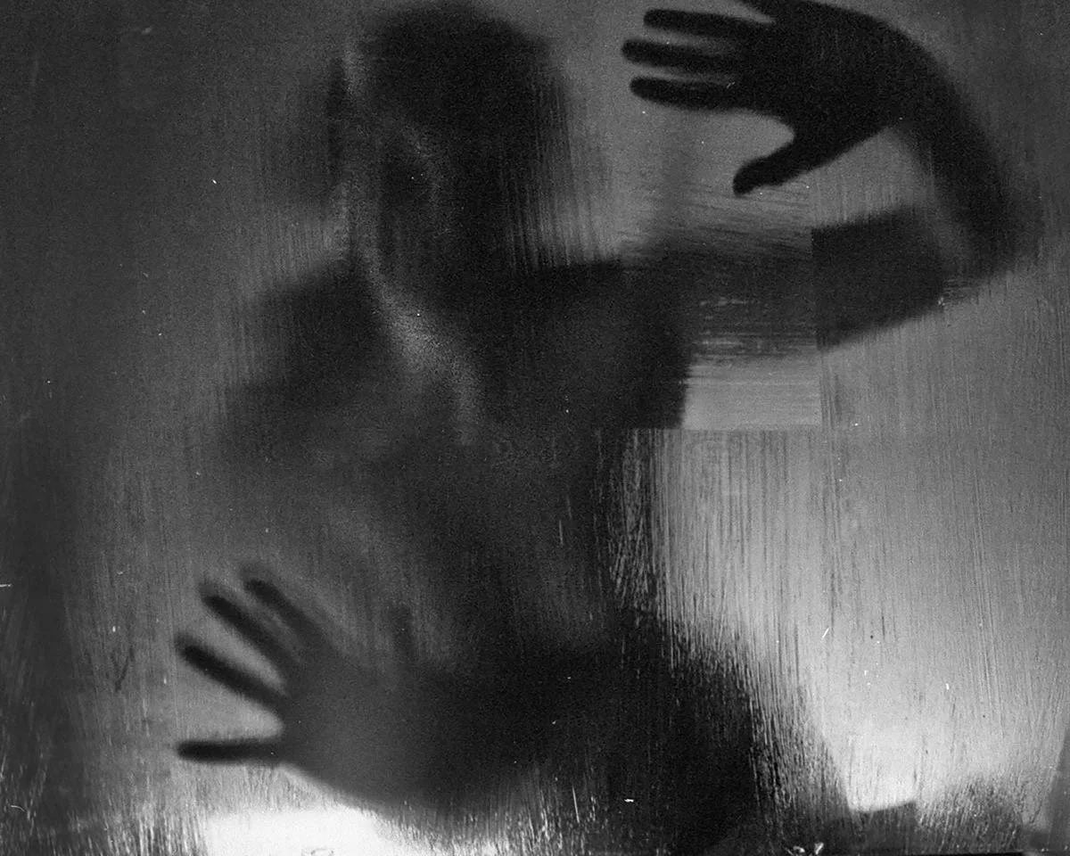 Silhouette of a person pressing their hands and face against a glass surface, creating a shadowed figure behind the glass.