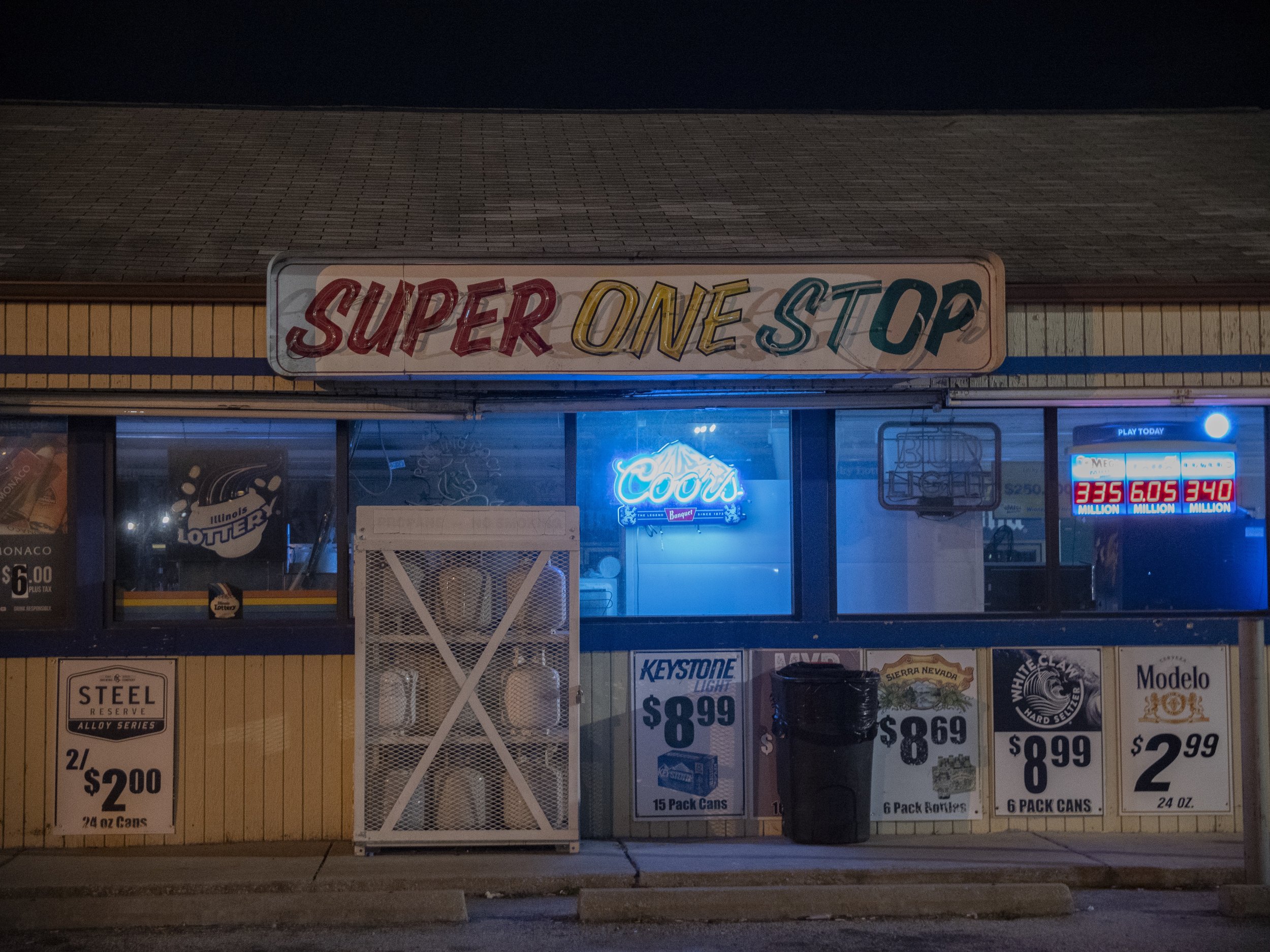 Front of a convenience store named "Super One Stop" illuminated with blue neon lights, showing drink advertisements, a basketball hoop, and a lottery sign.