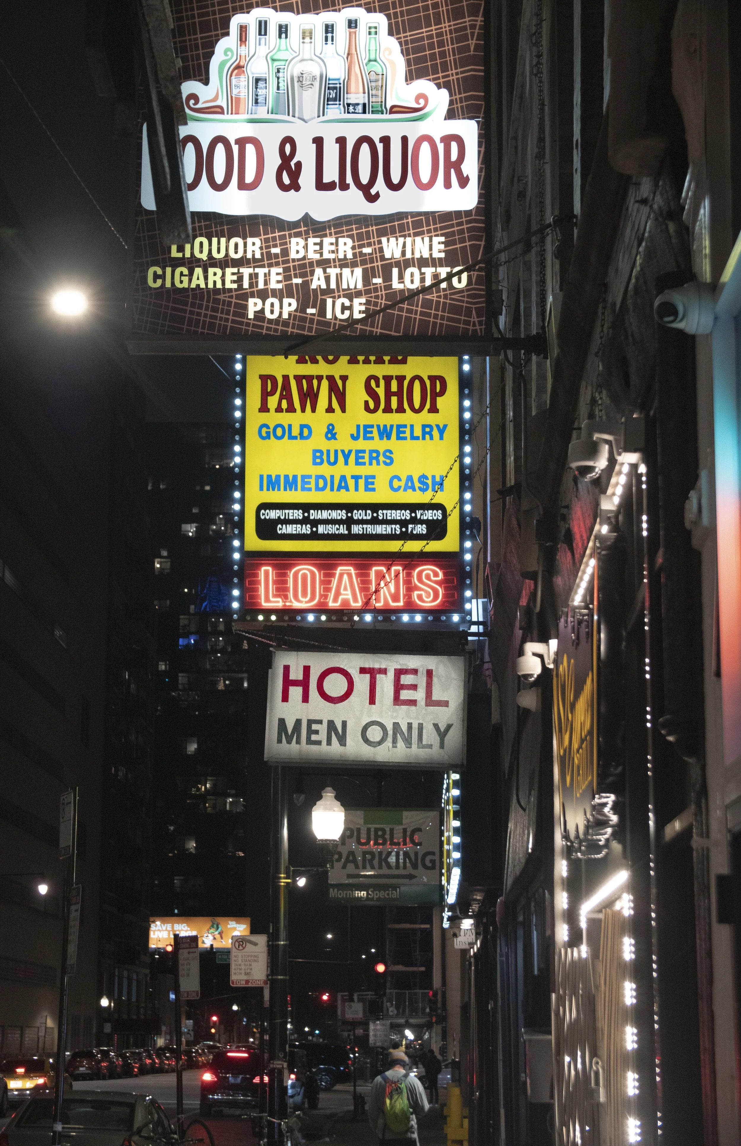 Nighttime city street with illuminated commercial signs, including a food and liquor store, pawn shop, hotel for men only, and parking sign, with pedestrians and cars in the background.