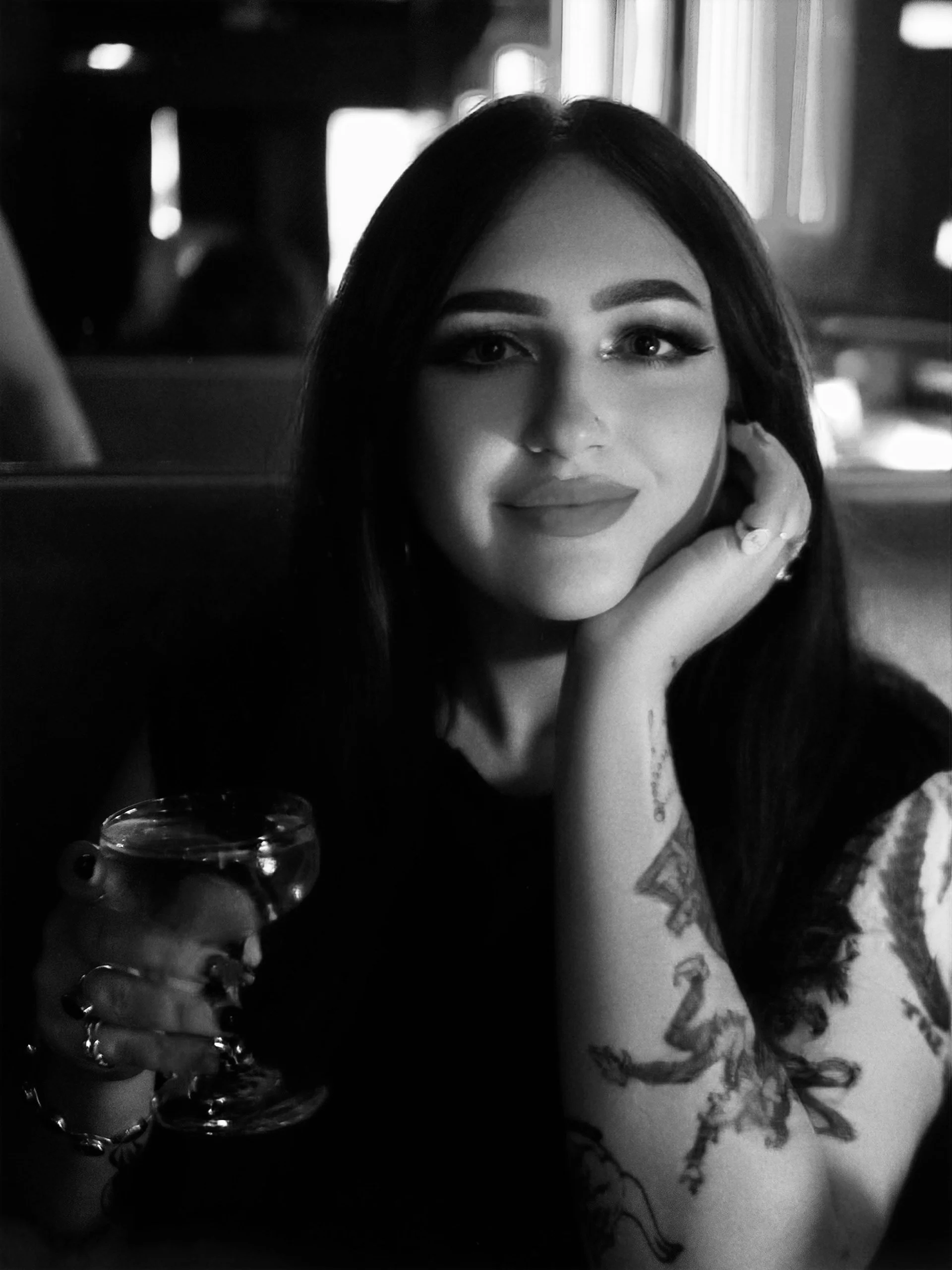 A woman with dark hair, makeup, and tattoos is sitting in a dimly lit setting, holding a glass of drink, smiling slightly, with her chin resting on her hand.