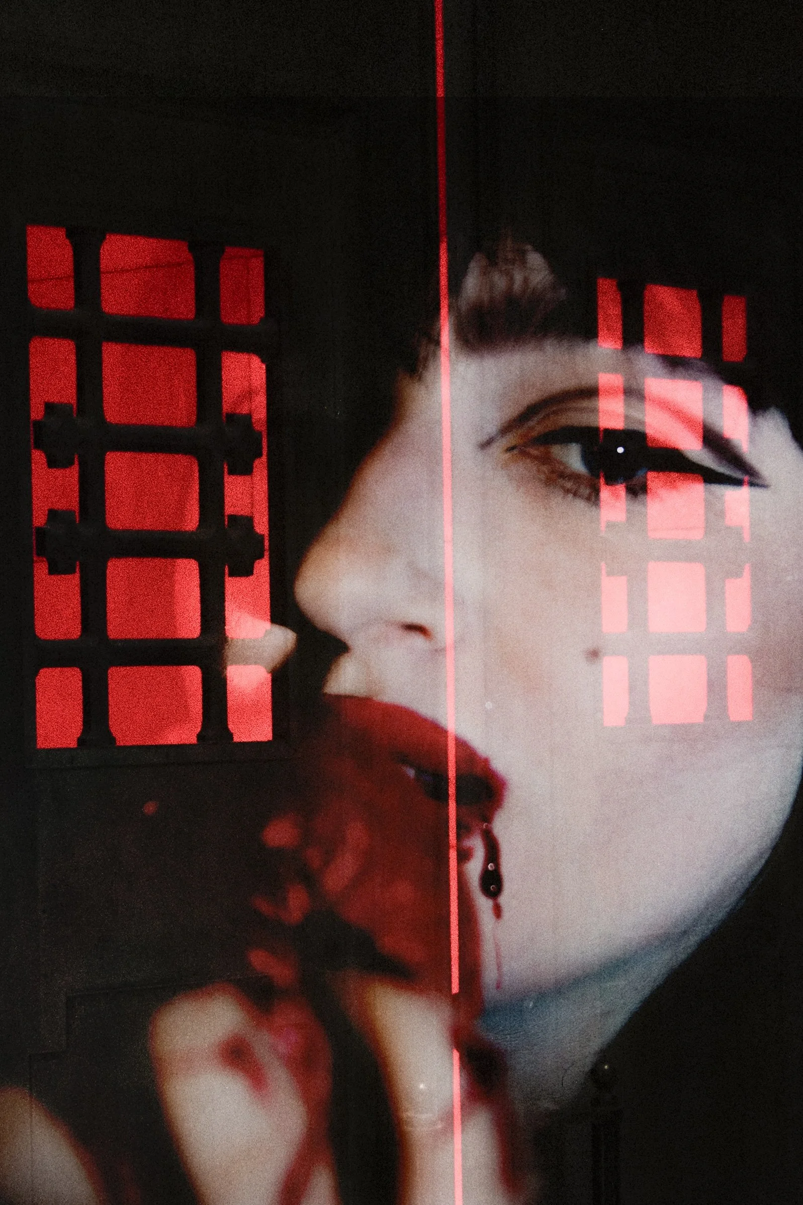 Close-up of a woman's face, with bold makeup, partially obscured by red light and grid-like reflections.