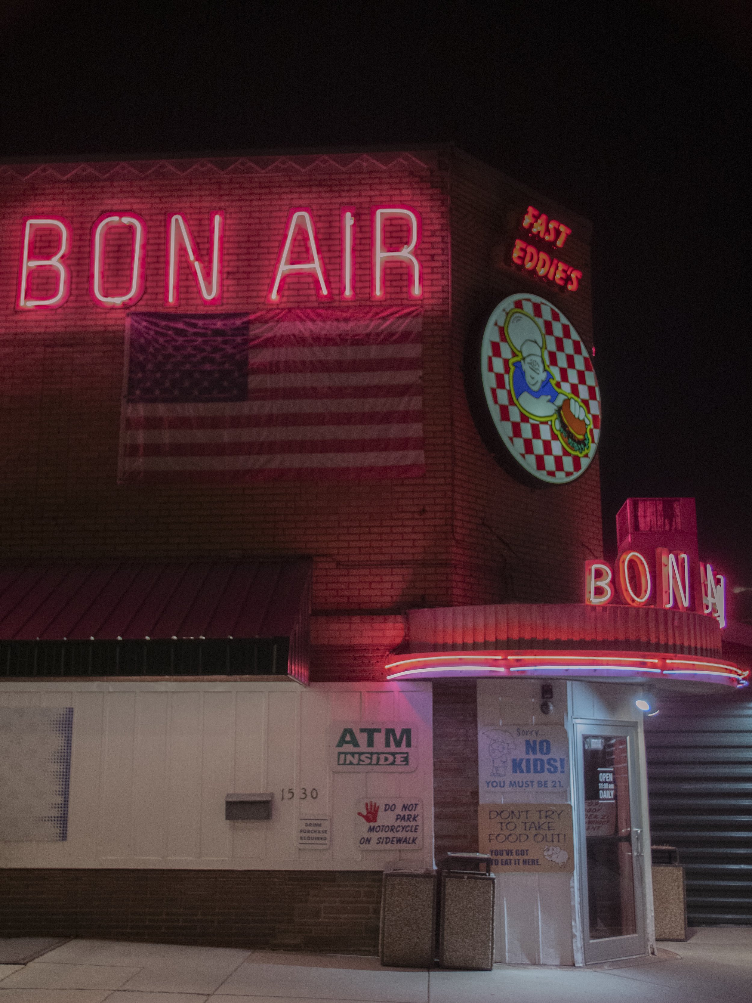 Nighttime exterior of Bon Air restaurant with pink neon signs, a large round logo with a chef, an American flag, and an ATM sign, neon lights around the entrance, and various posted signs near the door.