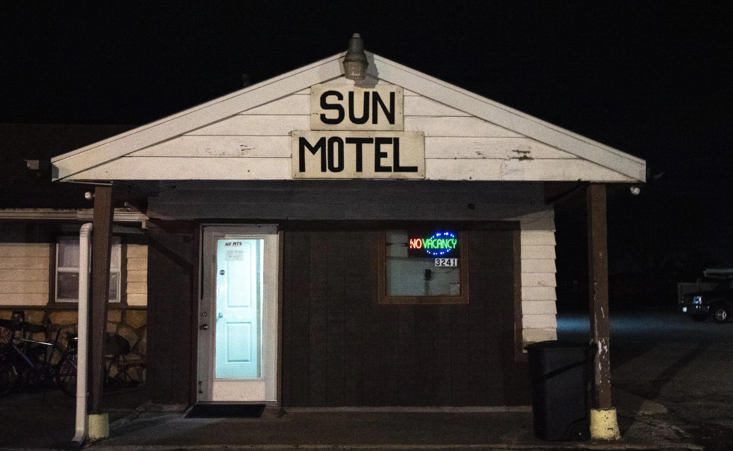 Night view of a small motel building with a sign reading 'Sun Motel', a closed white door, and a neon sign that says 'No Vacancy' in red and green colors.