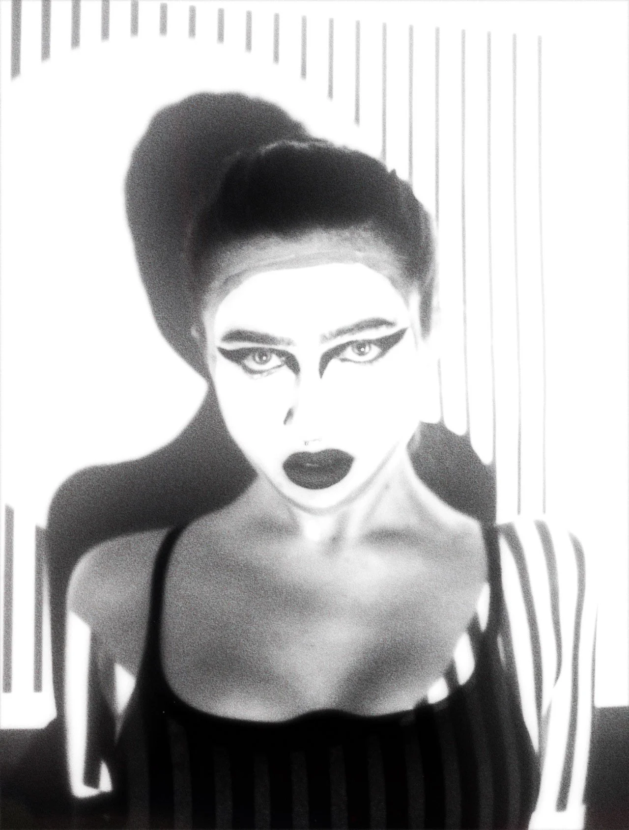 Black and white portrait of a woman with dramatic makeup, dark lipstick, and striped clothing, standing against a striped background.