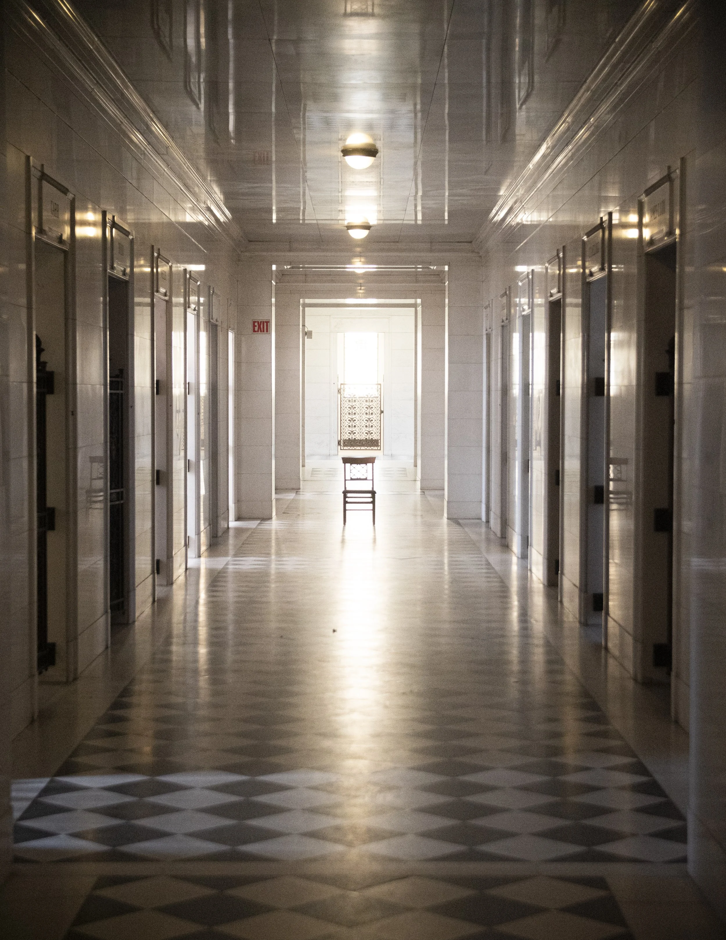 A symmetrical indoor corridor with elevators on both sides and a patterned floor leading to a bright room with a chair at the end.