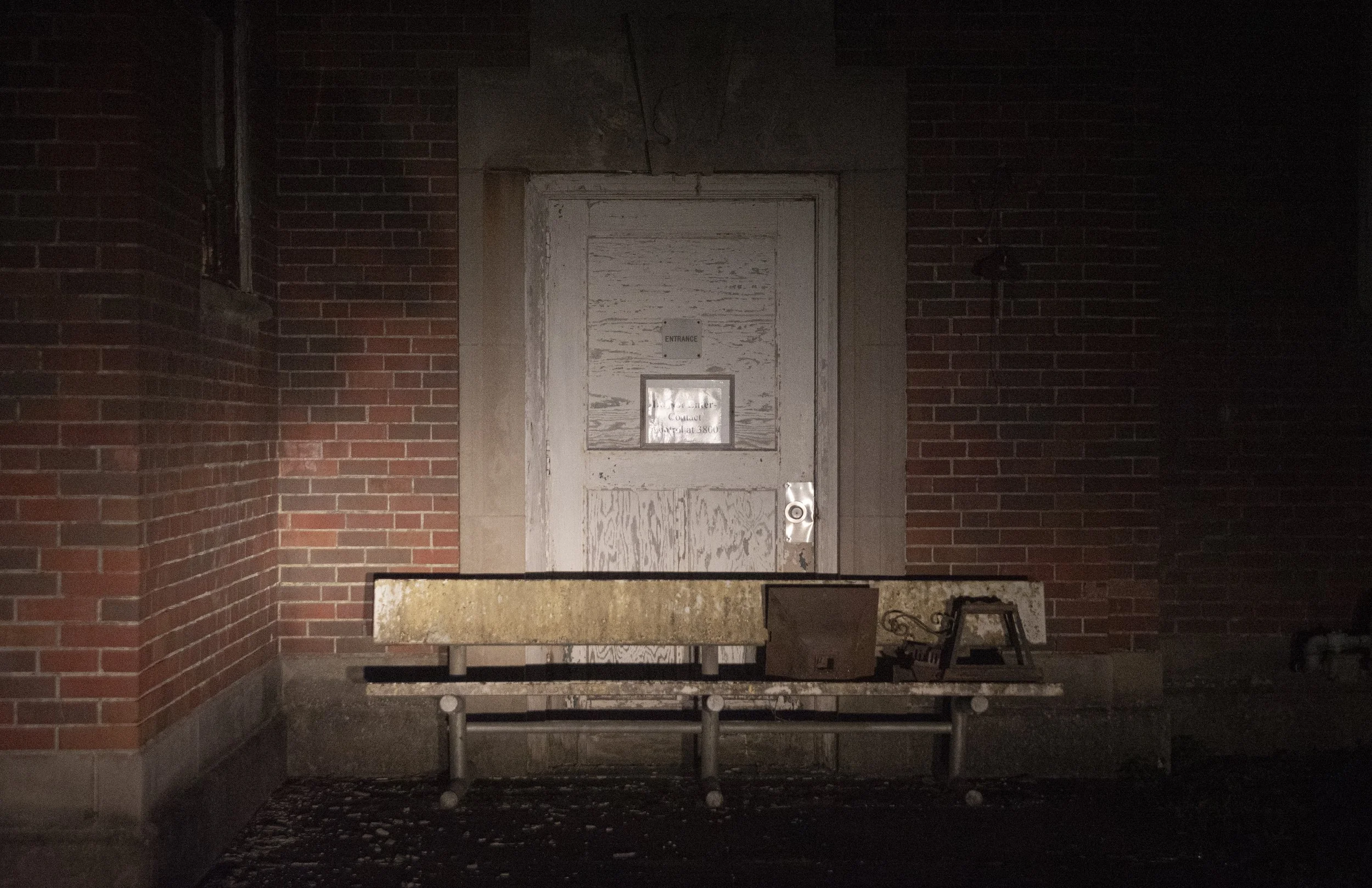 A boarded-up white door with a small window and a sign, set into a brick wall, with a long concrete and metal bench in front, some objects on the bench, and a dark surrounding.