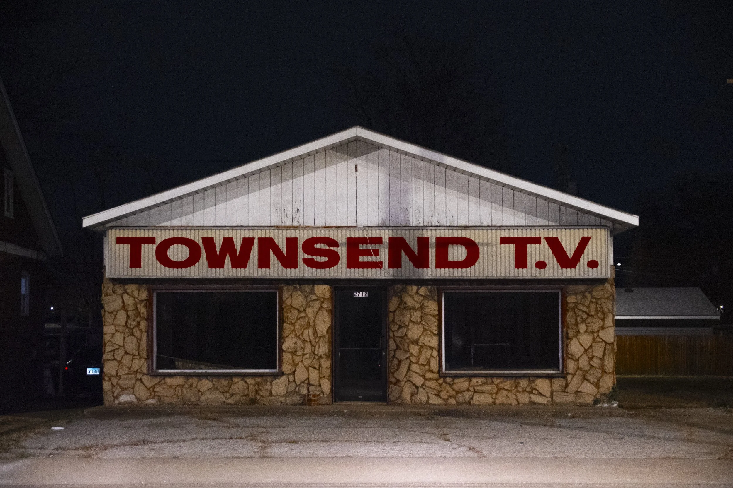 A small building with stone walls, two large dark windows, and a door in the center. The facade has a sign that reads 'TOWNSEND T.V.' in large red letters. It appears to be nighttime.