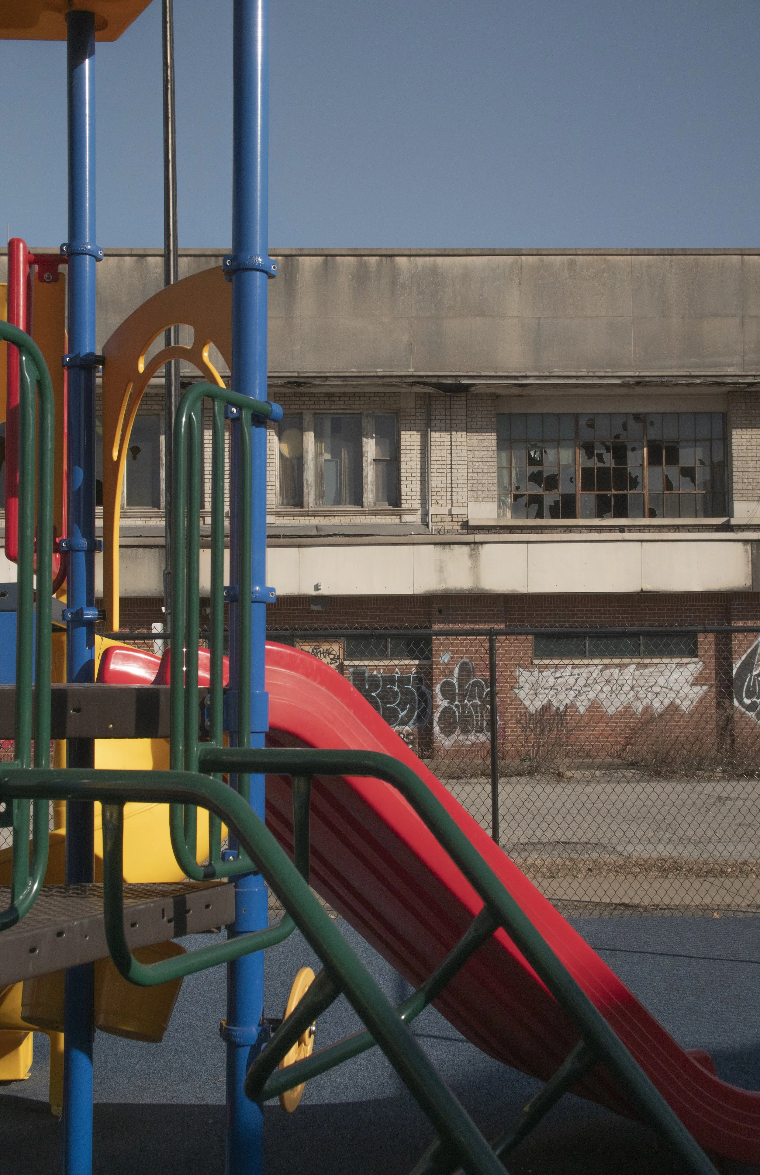 Colorful playground slide and climbing structure in an urban outdoor area, with an old building and graffiti-covered fence in the background.
