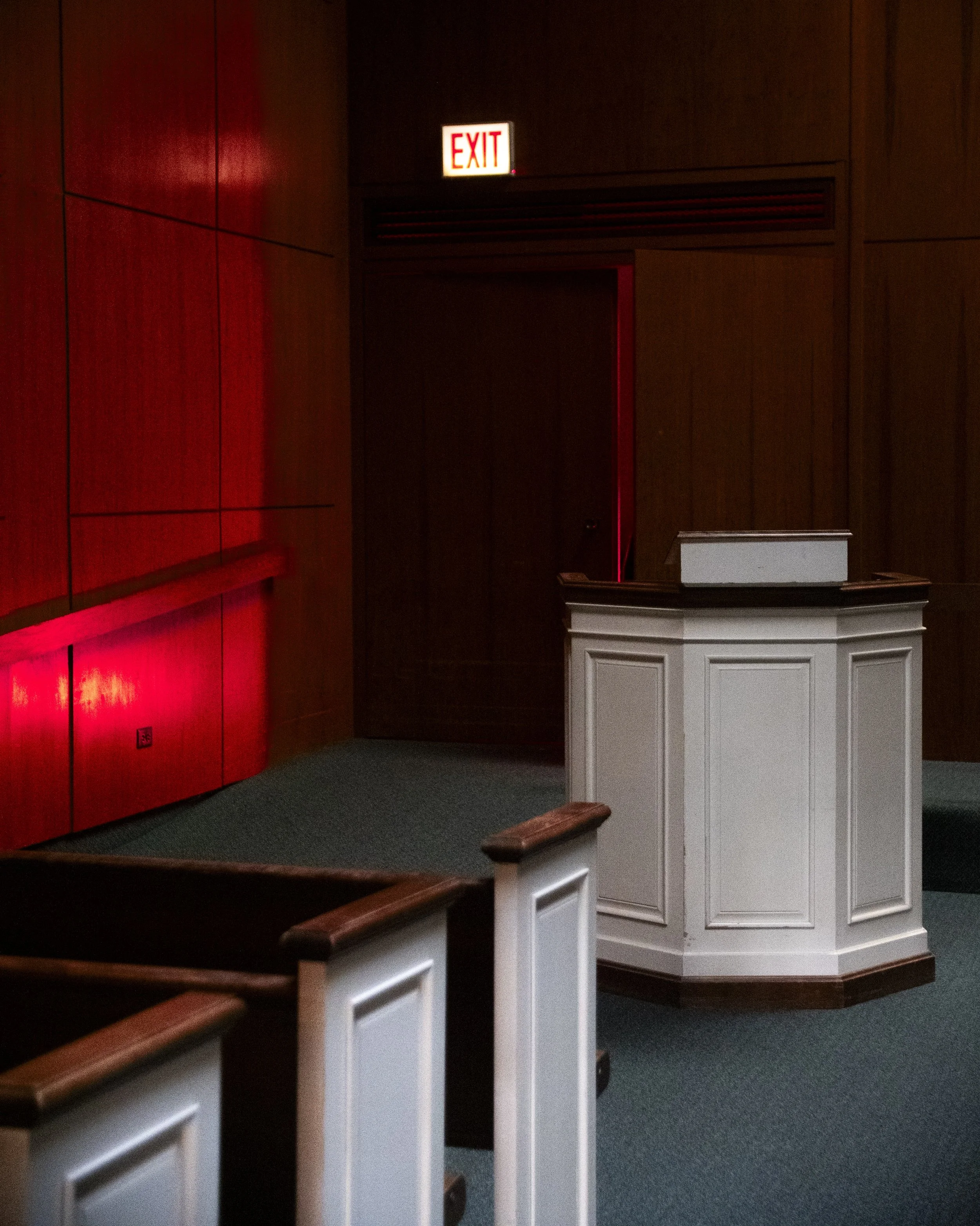 Empty courtroom with wooden walls, a white witness stand, and an exit sign above a door.