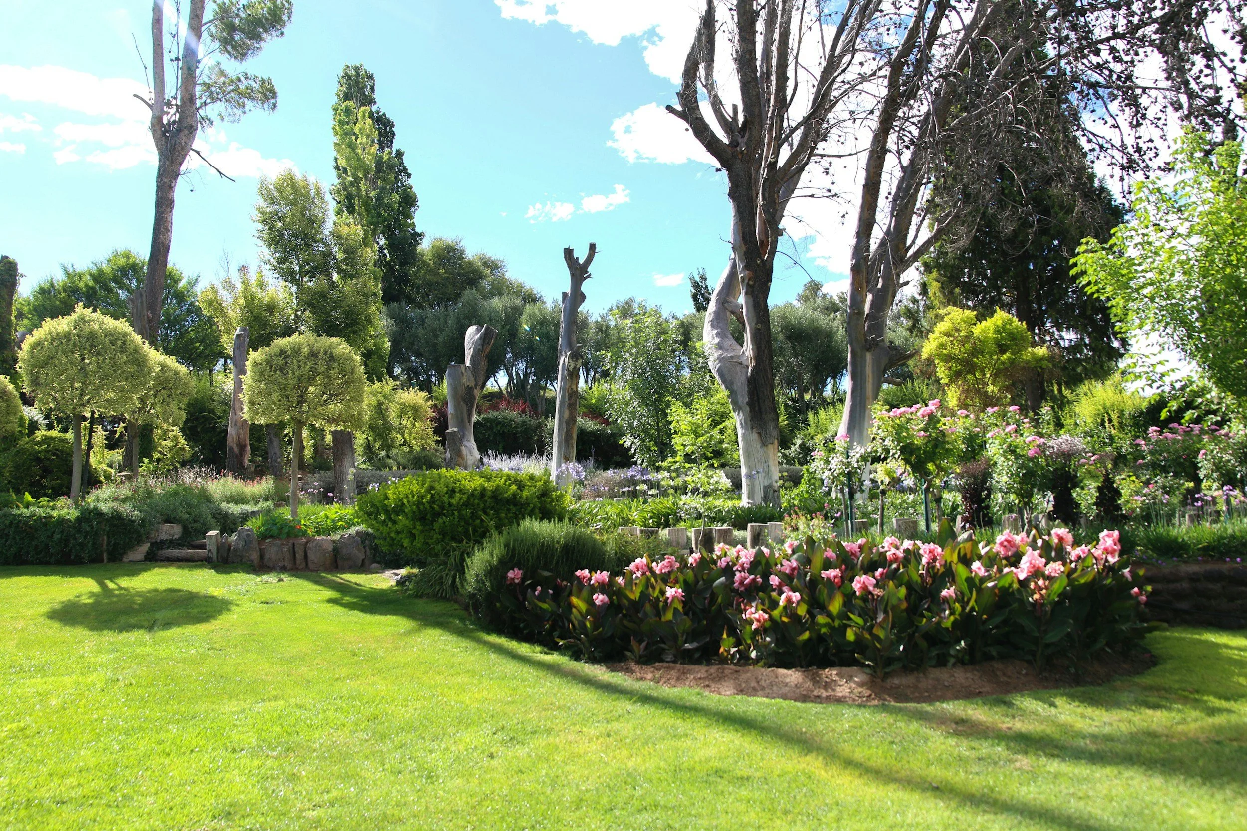 A lush garden with green grass, blooming pink and white flowers, and various trees, some with trimmed round canopies and others with tall, bare branches. The sky above is blue with scattered clouds.