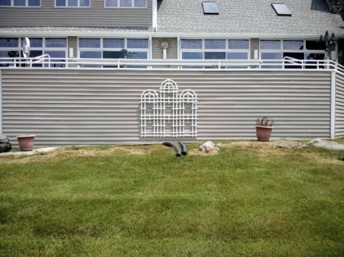 A residential backyard with green grass, two potted plants, a decorative white gate on a beige vinyl fence, and a house with large windows and a balcony in the background.