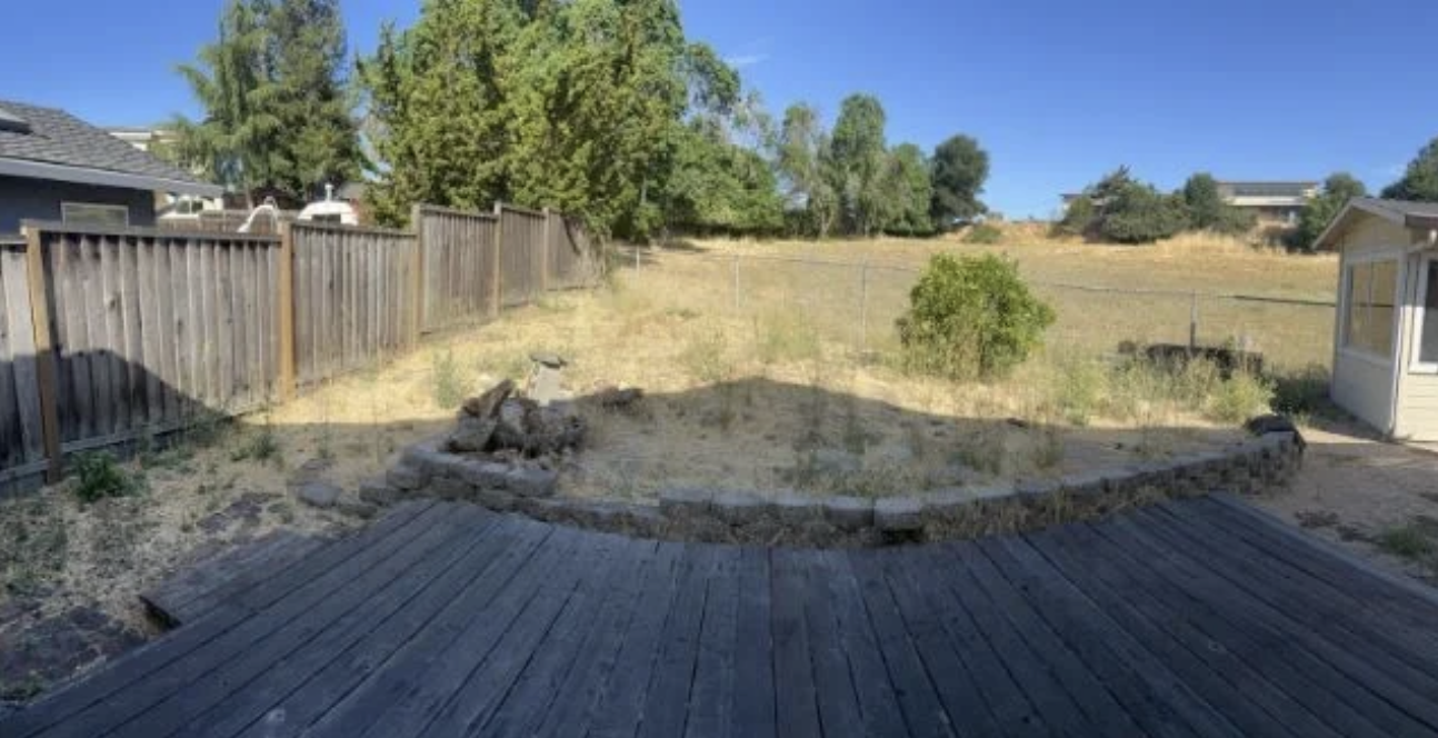 Backyard with a wooden deck, a partially raised garden bed with stone edges, a tall wooden fence, and a grassy, slightly sloped yard with trees and bushes under a clear blue sky.