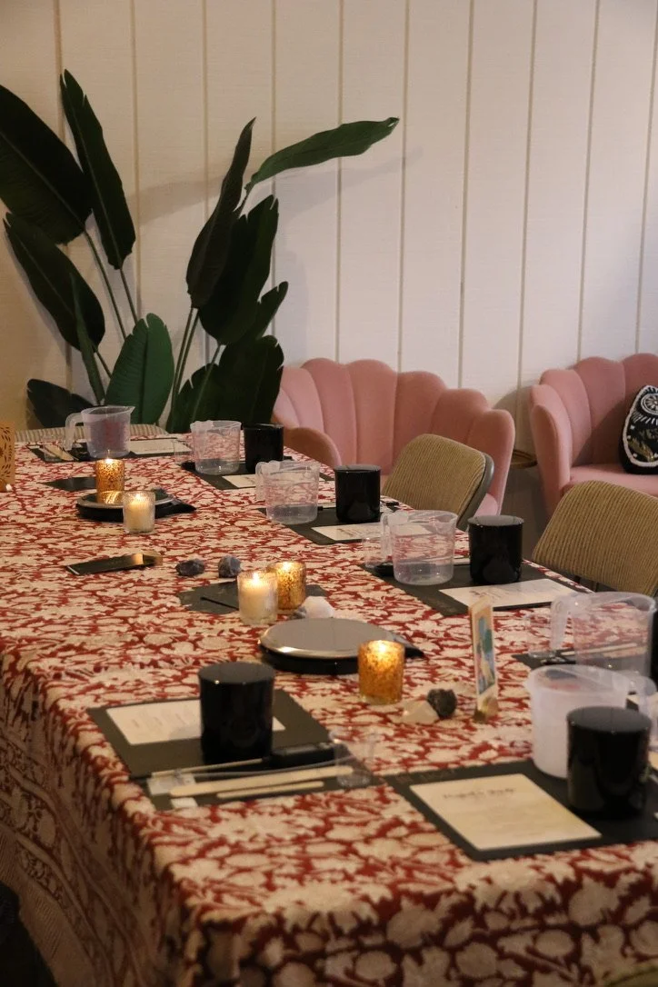 Set dining table with candles, black and clear cups, plates, and utensils in a cozy indoor setting, with pink cushioned chairs and tropical plants in the background.