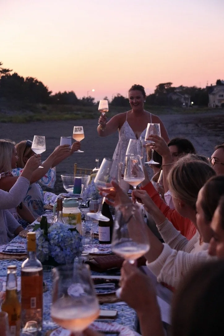 A group of friends enjoying an outdoor dinner on the beach at sunset, toasting with wine glasses.