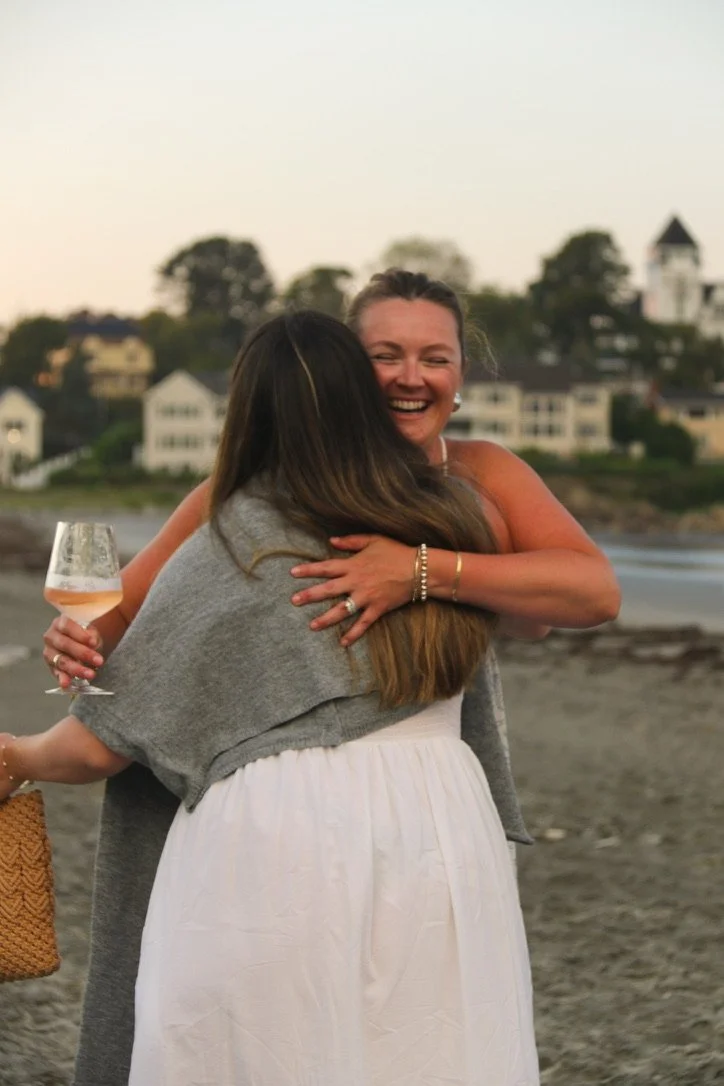 Two women hugging on a beach during sunset, one holding a glass of wine, with houses and trees in the background.