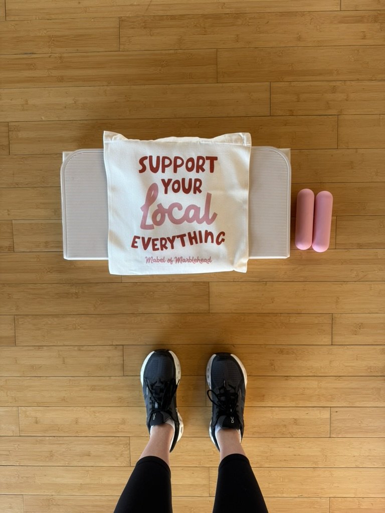 View of a person wearing black running shoes and black leggings standing on a wooden floor, with pink dumbbells, a white exercise step, and a tote bag with red and pink text that reads 'Support Your Local Everything' on the floor.