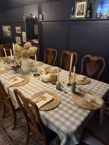 A dining table set for a meal with a checkered tablecloth, beige woven placemats, white plates with napkins, and tall white candles in candle holders. The table has a centerpiece of white and purple hydrangeas in a white vase, surrounded by small candles. There are eight wooden chairs around the table in a cozy, decorated dining room with framed pictures and decorative items on shelves in the background.