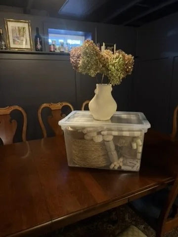A large plastic container holding a white vase with dried hydrangea flowers is placed on a wooden dining table. The background features a dark-colored wall with framed pictures and decorative bottles.
