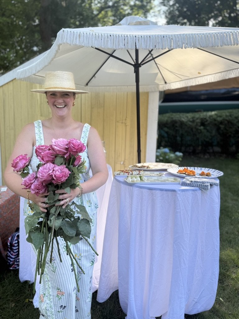 A woman smiling and holding a bouquet of pink roses outdoors, standing in front of a white table under a large white patio umbrella, with a yellow wooden fence and greenery in the background.