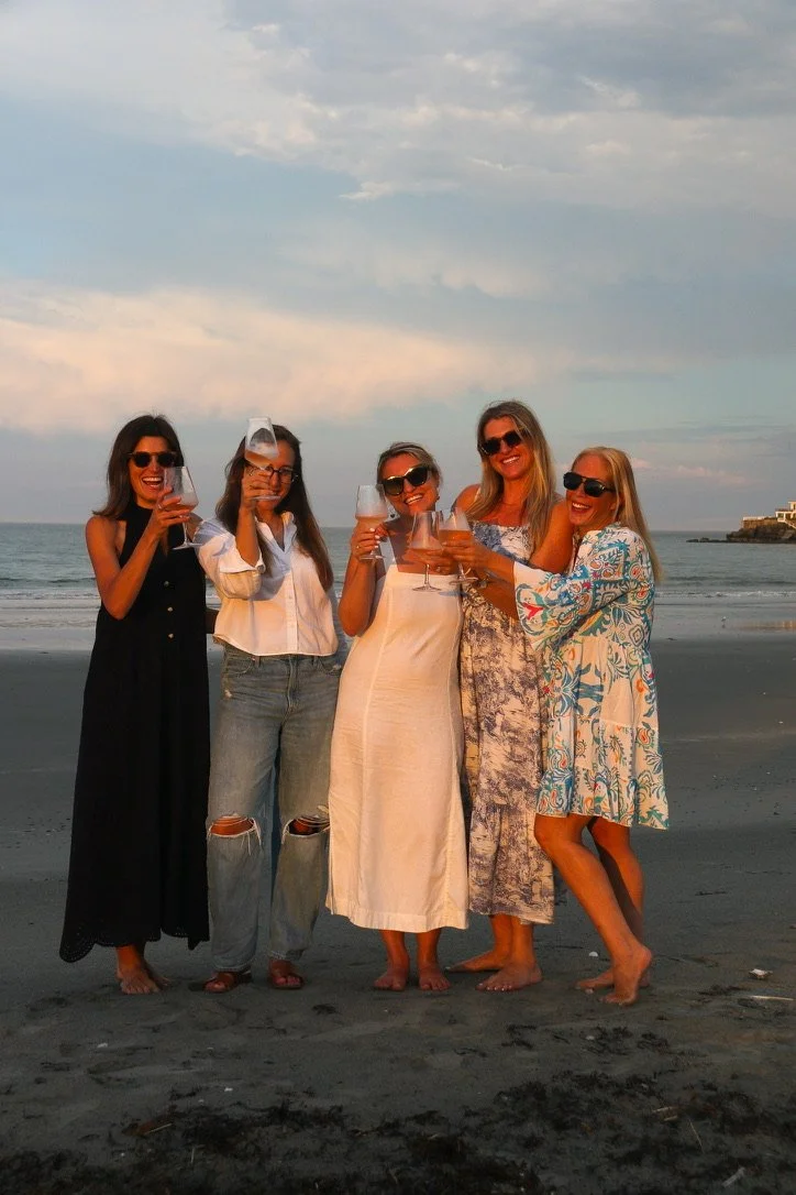 Five women in sundresses and sunglasses holding wine glasses, celebrating on the beach during sunset.