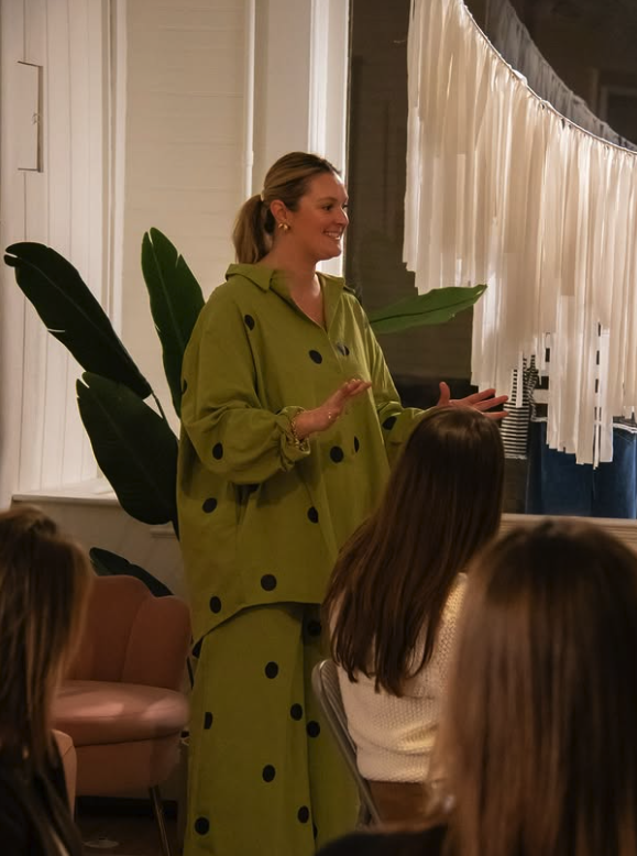 A woman in a green outfit with black polka dots speaking to an audience indoors, with a large green plant and white curtains in the background.